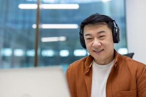 Asian man businessman in office using laptop for virtual meeting. Wearing headphones, smiling. Focus on digital communication, remote work, technology use. Bright modern setting, professional vibe. photo