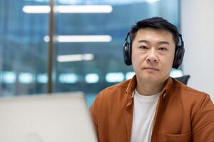 Asian man businessman wearing headphones in modern office environment focused on work. Seated at desk with laptop, exploring concepts of remote work, concentration, and communication. photo