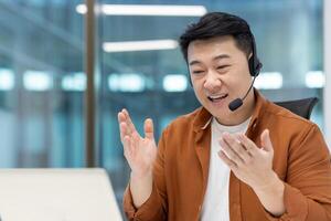 Mature Asian businessman with a headset communicating during an online meeting in a professional workspace, gesturing expressively, showcasing engagement and collaboration in virtual communication photo