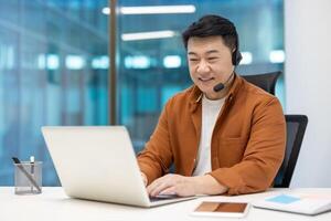 Asian businessman wearing headset working at laptop in modern office. Smiling, focused, multitasking. Smartphone on desk enhances connectivity. Represents professionalism, communication, success. photo
