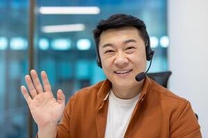 Smiling asian businessman using headset for call. Friendly professional waving hand, engaging in communication, demonstrating modern tech use in office environment. photo