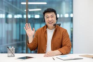 Asian man wearing a headset and smiling waving in office. Represents communication, technology, interaction. Ideal for concepts of customer service, online conferencing, or professional support. photo