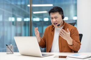 Angry man with headset talking while sitting at laptop inside office, dissatisfied employee yelling at interlocutor, unable to communicate with client. photo
