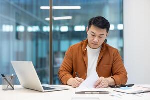 Asian man working on papers at a desk in a modern office environment, emphasizing productivity and focus. photo
