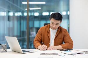 Asian man in a professional office environment writing notes while working with documents and a laptop, showcasing dedication and focus. Concept of business routine, office work, and productivity. photo