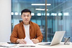 Portrait of serious successful financier investor. Man behind paperwork looking at camera with concentration. Businessman working with laptop and documents inside office. photo