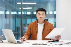 Mature Asian man reviewing documents and working on a laptop at a desk in a modern office. Represents productivity, professionalism, and technology in a business setting. photo