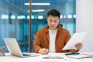 Asian businessman sits at desk working with documents, demonstrating focus and productivity. Man holds pen, works with laptop, showcasing professional business setting. Ideal office themed content. photo