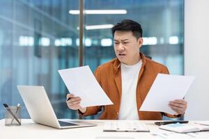 Asian man analyzing financial paperwork at an office, expressing dissatisfaction with a financial report. Businessman working on taxes, reviewing deductions, and evaluating financial statements photo