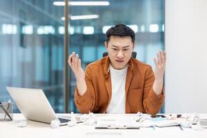 Asian man experiencing frustration and stress while working at a cluttered desk in an office. Overwhelmed by workload, he sits surrounded by crumpled papers and tries to manage challenges. photo