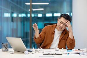 Asian businessman showing frustration while sitting at office desk with laptop and crumpled paper. Man holding head, experiencing work stress and fatigue. Office setting. Workplace challenges. photo