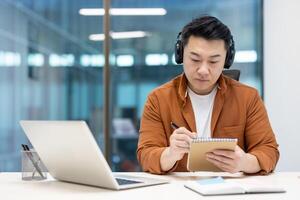 Asian man with headphones and notepad watching educational course webinar. Man recording data listening teacher conversation. Businessman using laptop for call and remote online meeting photo