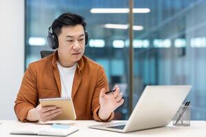 Asian man wearing headphones using a laptop for an online meeting, dressed casually in an office setting. He holds a notepad in one hand and writes with a pen, showing focus and productivity. photo