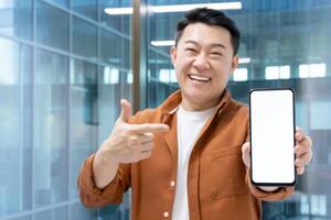 Asian man mature businessman happily showcasing a blank smartphone screen in modern office setting. Perfect image for concepts of technology, communication, and positivity photo