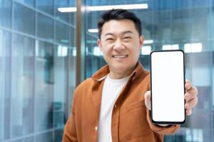 Asian businessman showing phone screen to camera, man at workplace with smartphone smiling and looking at camera, working inside office. photo