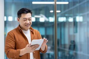 Asian man interacting with a tablet in an office. Demonstrates technology use and communication in a professional setting, reflecting productivity and modern work practices. photo