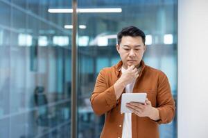 Asian man focusing on a task using a digital tablet in an office. His thoughtful expression suggests active problem-solving, concentration, and workplace productivity within a professional setting. photo