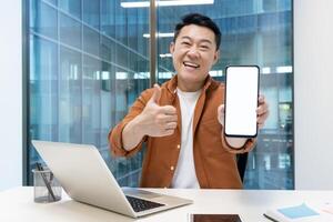 Asian man presenting and showing a smartphone while sitting at a desk in an office. The individual appears confident and joyful, highlighting the blank screen for any customizable display photo