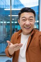 Asian businessman having call using phone in office, wearing casual attire. Expression of happiness and connection in professional setting. Gesture of communication displays confidence. photo