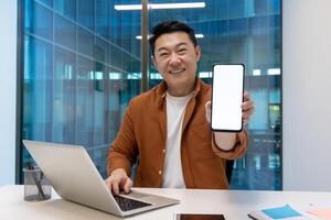 Asian businessman in office holds smartphone with blank screen, smiles confidently. Works on laptop at desk, displaying technology professionalism, innovative spirit. Modern workspace vibes. photo