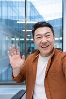 call, Asian man looking into phone camera and talking to colleagues using smartphone app. Businessman at workplace inside office. photo