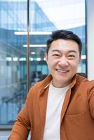 An Asian mature businessman takes a selfie call with his phone, smiling confidently in a modern office environment, expressing professionalism and a positive demeanor. photo