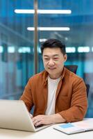 Asian businessman smiling while working on laptop in office, showing productivity, concentration, and focus. Engaged in tasks, embodying efficiency and modern workplace innovations. photo