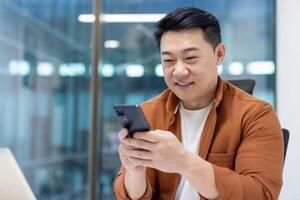 Close-up of successful Asian man using phone, smiling man typing text message, satisfied businessman in casual shirt inside office. photo