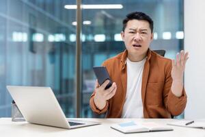 Asian man displaying frustration in a modern office, seated at a desk with a smartphone in hand, showing emotions of concern and confusion, surrounded by work materials and professional tools. photo