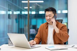 Asian businessman smiling while multitasking with phone and laptop in modern office setting, demonstrating effective communication and productivity. Concept of technology in business environment. photo
