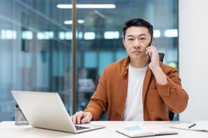 Portrait of a businessman inside the office at the workplace, a man is talking on the phone seriously and looking at the camera with concentration. Asian man at work with a laptop. photo