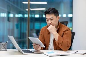 Asian businessman in office focusing on tablet with laptop, reflecting on strategy. Casual reflects modern work environment. Tech-savvy professional analyzing data, considering business decisions. photo