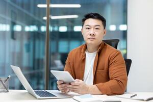 Portrait of serious and concentrated man at workplace inside office. Asian man looking at camera while holding tablet computer. Experienced office worker programmer developer. photo
