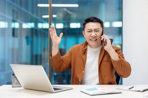 Asian businessman expressing frustration during business call in modern office. Man seated at desk with laptop, using phone, gesturing in conversation, showcasing emotion and communication. photo