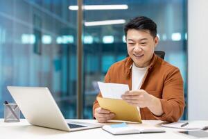 Happy Asian businessman receiving exciting mail at office, grinning while reading message. Professional workplace setting, laptop and documents present. Expression of success and enjoyment at work. photo