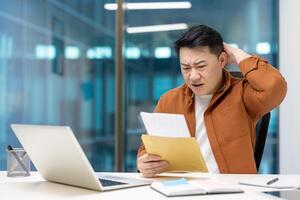 Asian businessman looks surprised receiving mail in office. Expression of concern indicates unexpected or bad news. Enveloped mail held in hand, open laptop on desk. photo