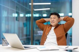 Asian businessman smiling while taking break in office. Man sitting back, relaxed, looking satisfied. Laptop and documents on desk suggest workspace. Concept of leisure, balance. photo