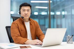 Portrait of an Asian worker with a headset and a laptop inside the office. The man is looking at the camera seriously and concentrated, a support and remote consulting worker. photo
