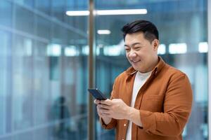 Successful Asian businessman standing near the window with phone in hands. Man using application on smartphone, typing message and browsing social media inside office at workplace. photo