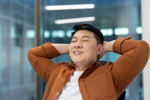 Asian man, dressed casually, leaning back with hands behind his head in modern workspace, expressing relaxation and satisfaction with a joyful smile. photo