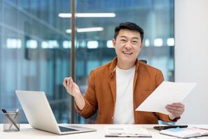 Portrait of happy and successful Asian financier investor. Man smiling and looking at camera, holding report in hands, satisfied with results achieved at workplace, working inside office with laptop. photo