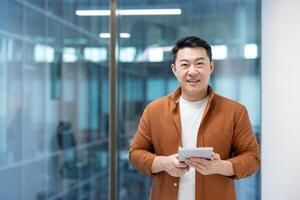 Portrait of successful Asian man inside office. Businessman smiling and looking at camera while holding tablet computer. Programmer developer at workplace near window. photo
