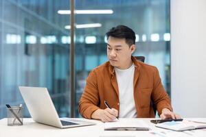 Serious confident Asian financier working on paperwork inside office, businessman with calculator calculating and filling out financial reports and tax forms inside office with laptop. photo