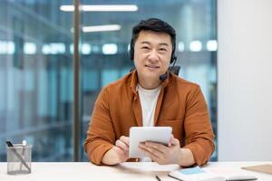 Portrait of successful Asian worker with headset and tablet computer in hands. Man smiling and looking at camera, working inside office at workplace. Online support service worker. photo