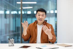 Webcam view, call, Asian man looking at camera, smiling and communicating with colleagues and partners remotely. Successful businessman working inside office at workplace. photo