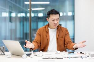 Disappointed and upset asian man among pieces of torn paper at workplace, financier dissatisfied with performance. Businessman angry and sad with bad report. photo