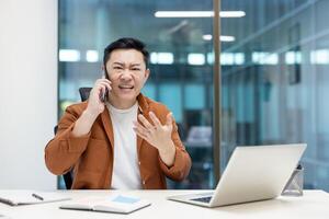 Angry asian man talking on the phone inside office at workplace shouting. Businessman looking at camera dissatisfied, working with laptop. photo