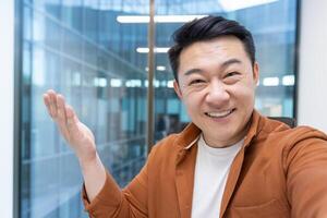 Happy and successful Asian man talking using call app on phone. Man working inside office at workplace, looking at camera. photo