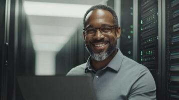 Man with glasses and beard smiles while using laptop in a professional server room setup photo