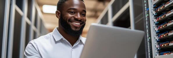 Man smiles while using a laptop in a busy data center with numerous server racks around photo